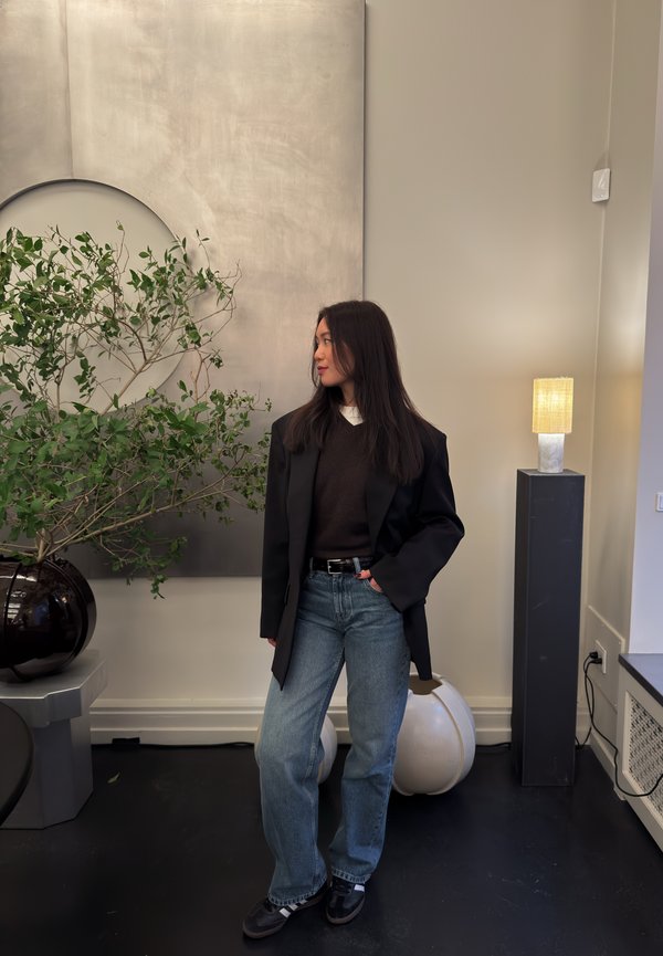 Mujer con blazer negro y jeans de pie en el interior, mirando hacia la izquierda, cerca de una planta grande en una maceta oscura y una lámpara de pie sobre un pedestal alto.