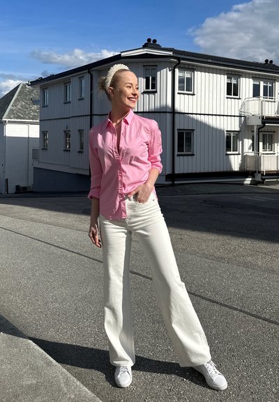 Mujer con una camisa rosa de botones y pantalones anchos blancos se encuentra en una calle pavimentada. Lleva zapatillas blancas y una diadema de perlas.