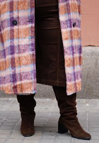 Άτομο που φοράει παλτό με μοβ, πορτοκαλί και λευκό καρό, σκουρό καφέ φούστα από κορντόνι και σκουρό καφέ σουέντ μπότες μέχρι το γόνατο σε πλακόστρωτο δρόμο.