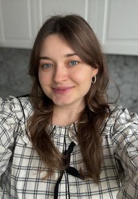 Jeune femme aux longs cheveux bruns et aux yeux bleus, portant des boucles d'oreilles anneaux et un chemisier à carreaux avec un ruban noir, souriante à l'intérieur.