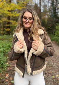 Smeđi jakna od ovčje kože s kremastim umetkom od umjetnog krzna na ovratniku i manžetama, sa zatvaračem sprijeda koji prikazuje vidljive detalje šivanja. Svijetla pozadina s lišćem.