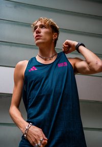 Fiatal férfi kék BMW Berlin Maraton trikóban, karját megfeszítve, nyakláncot, gyűrűket, karkötőt és karórát viselve, szürke vízszintes panel háttér előtt.