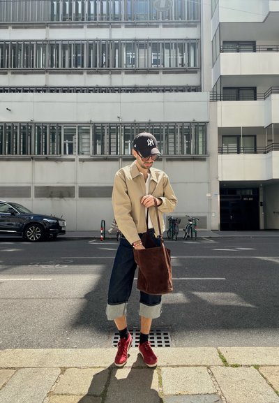 Bolso tote de ante marrón, chaqueta beige, pantalones de mezclilla oscuros y cortos, zapatillas rojas, gafas de sol y gorra. Entorno urbano con edificios.