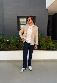 Bežs teksturēts krekls ar pogām uz baltā T-kreklā, apvienots ar tumšām biksēm un pelēkiem sporta apaviem; stāvot priekšā zaļumiem un pelēkai siena.