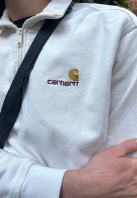 Sweatshirt branco com fecho em zíper e punhos canelados, apresentando um logótipo bordado da Carhartt em dourado e bordô na área do peito esquerdo.