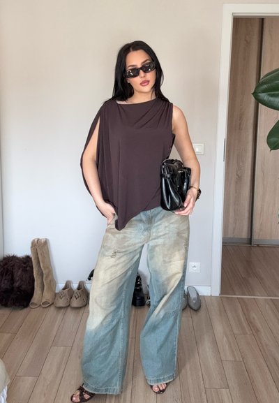 Mujer con top marrón sin mangas, de gran tamaño, jeans azules claros y anchos, gafas de sol negras, sosteniendo un bolso negro en una habitación con suelo de madera y zapatos al fondo.