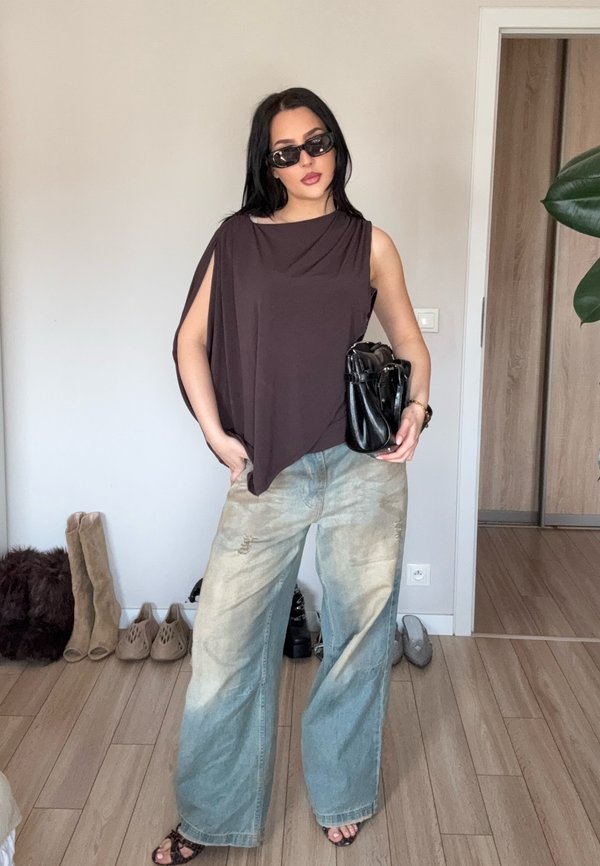 Femme portant un haut marron sans manches surdimensionné, un jean large bleu clair, des lunettes de soleil noires, tenant un sac à main noir dans une pièce avec un sol en bois et des chaussures en arrière-plan.