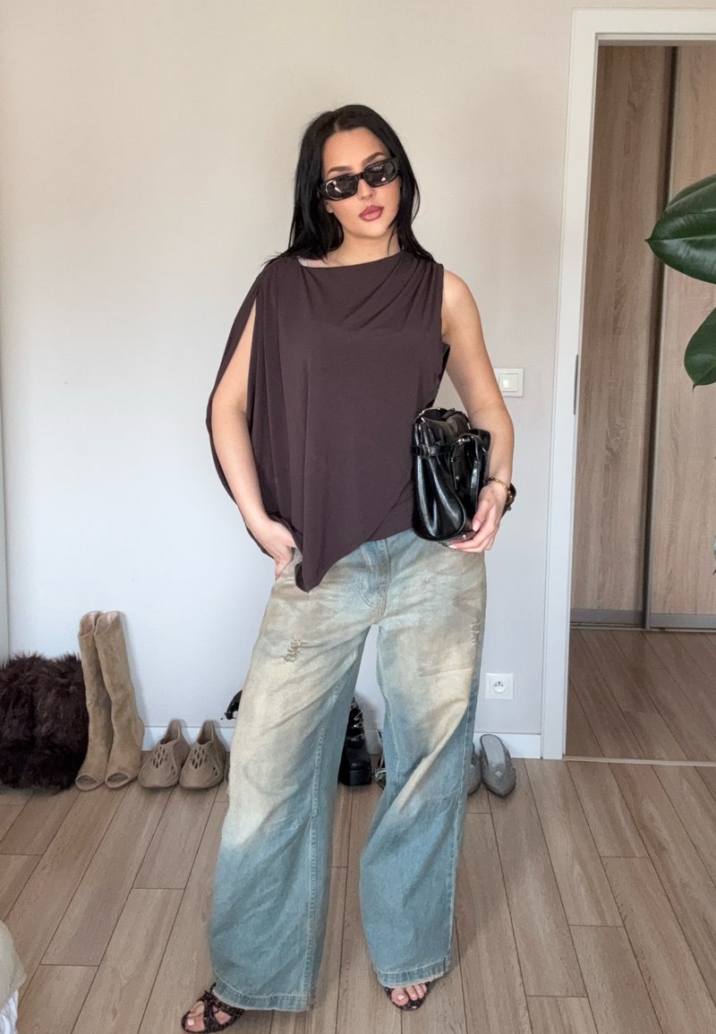 Femme portant un haut marron sans manches oversize, un jean large bleu clair, des lunettes de soleil noires, tenant un sac à main noir dans une pièce avec un sol en bois et des chaussures en arrière-plan.