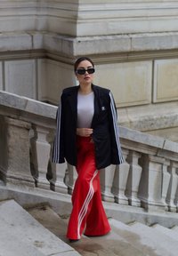 Blazer noir avec des rayures blanches, haut blanc, et pantalon large rouge avec trois rayures blanches ; lunettes de soleil noires et boucles d'oreilles.