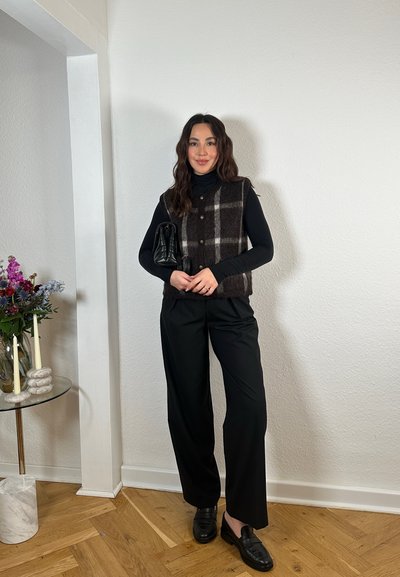 Chaleco de lana de cuadros marrones y blancos sobre un suéter de cuello alto negro, combinado con pantalones negros y mocasines. Sosteniendo un bolso negro texturizado.