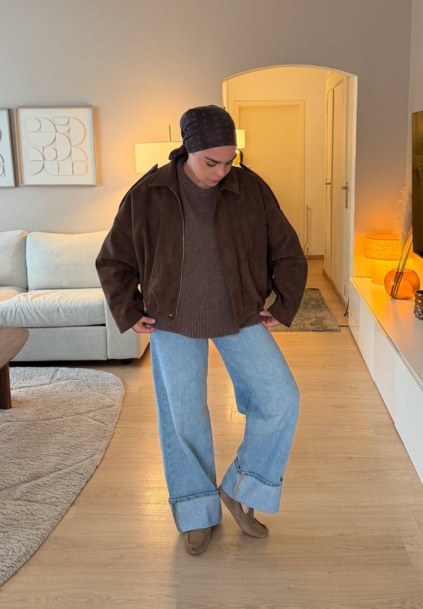 Une personne porte une veste brune, un pull gris, un jean bleu clair aux ourlets retroussés et des chaussures brunes dans un salon baigné d’une lumière chaleureuse.
