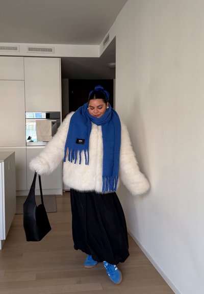Chaqueta de piel sintética blanca, bufanda azul con flecos, falda negra, zapatillas azules y bolso negro. Ambiente interior con suelo de madera clara.