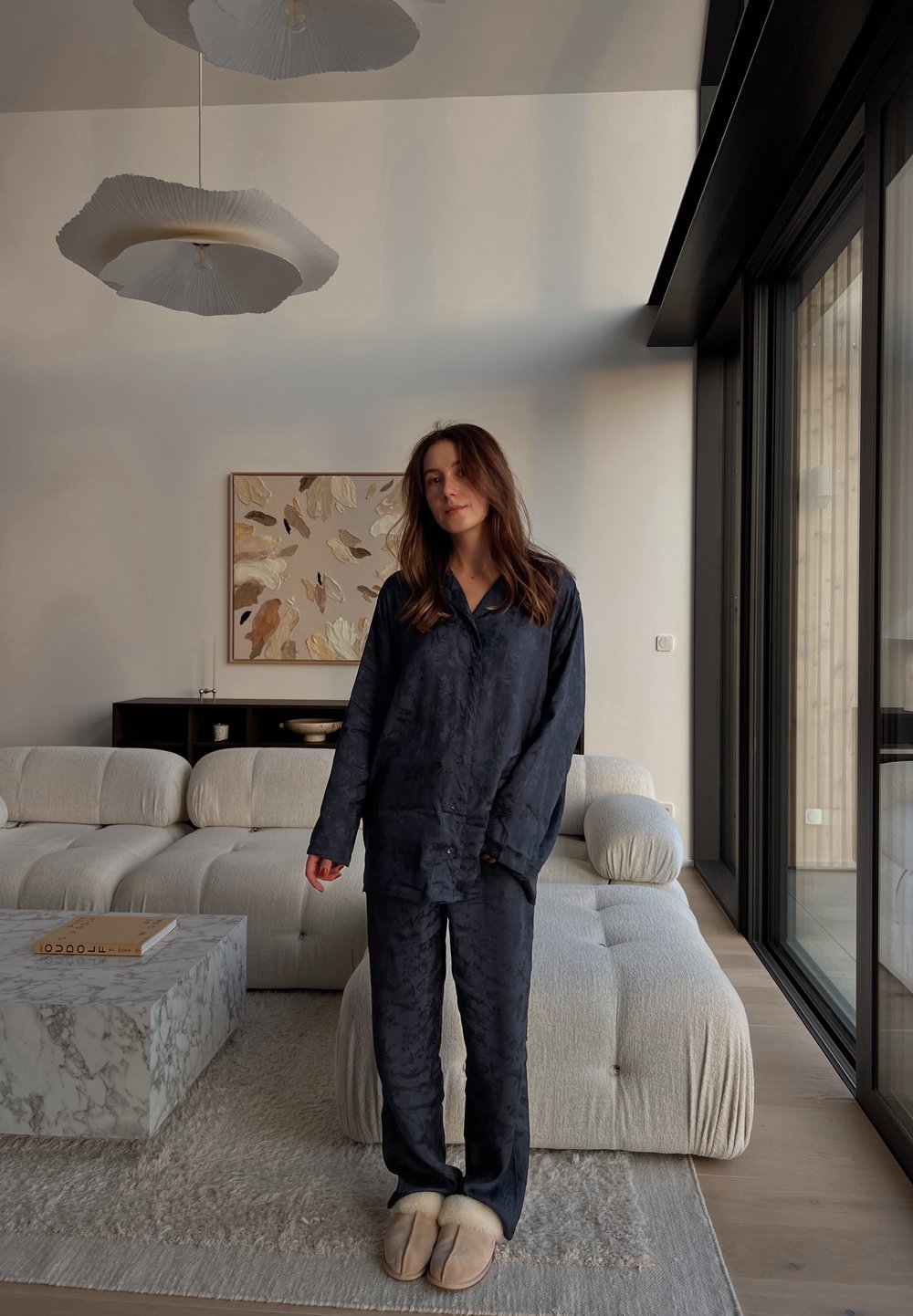 Satenast komplet pižame mornarsko modre barve z rožnatim vzorcem, dolgimi rokavi in hlačami. Mehki bež copati. Svetel teksturiran kavč in miza iz marmorja.