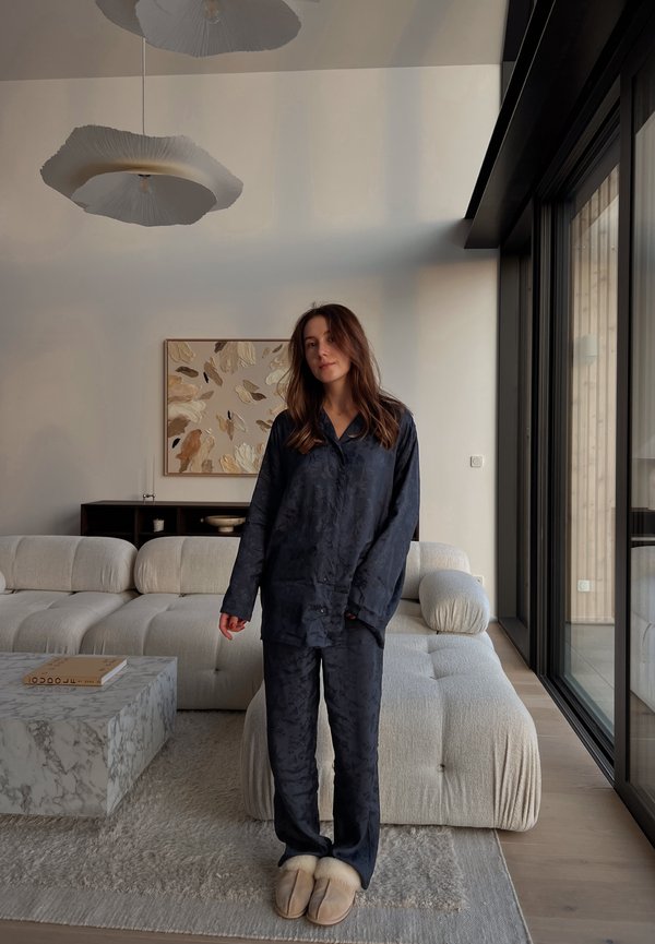 Ensemble de pyjama en satin bleu marine avec motif floral, manches longues et pantalon. Chaussons en peluche beige. Canapé texturé de couleur claire et table en marbre.