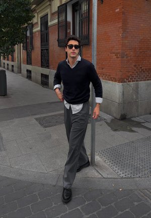 Joven con gafas de sol, suéter negro y pantalones grises caminando por la acera de una ciudad cerca de una esquina de un edificio de ladrillo con rejas metálicas en las ventanas.