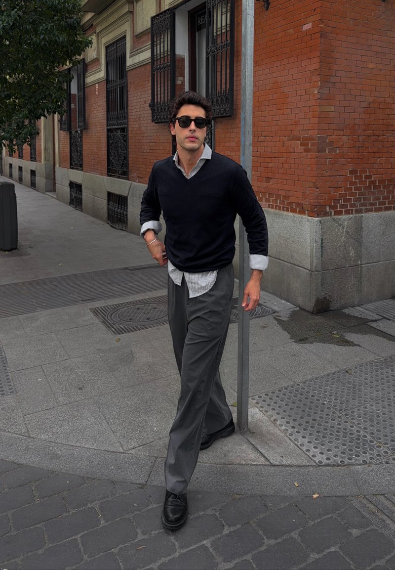 Jeune homme portant des lunettes de soleil, un pull noir et un pantalon gris, marchant sur un trottoir en ville près d’un coin de bâtiment en briques avec des grilles métalliques aux fenêtres.