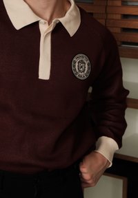 Burgundijas džemperis ar beidzīgu apkakli un uzlīmi, kas attēlo "Aizliegts Berlīnē". Struktūrēts audums, ribota aproce un pogu aizdare pie kakla.