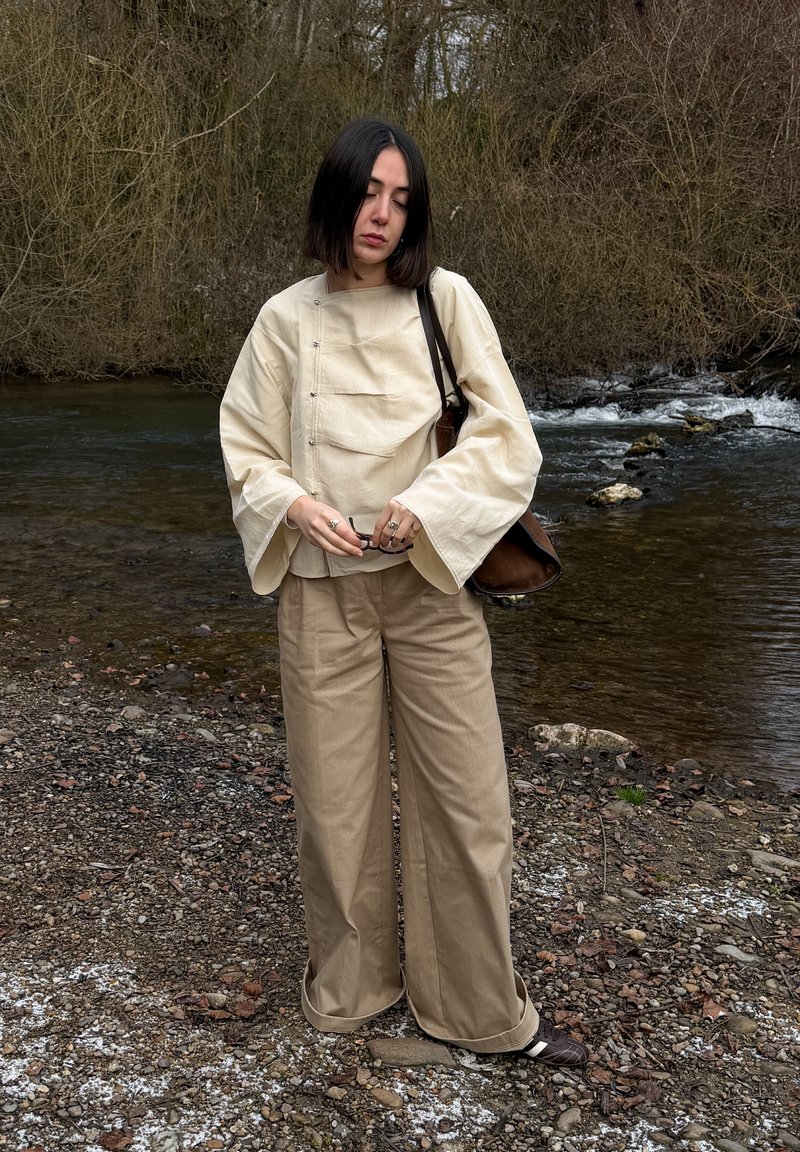 Woman with black bob hair wearing beige wide pants and loose cream top, standing on rocky riverbank with brown trees and flowing water background.