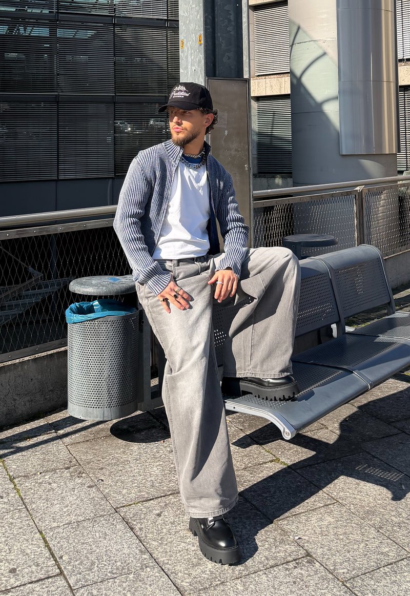 Man wearing black cap, white shirt, ribbed cardigan, wide grey trousers and black boots sits on outdoor bench near a bin in an urban setting.
