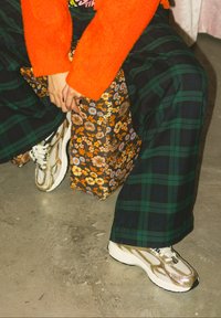 Personne portant un pantalon à carreaux verts, une veste orange duveteuse, une jupe fleurie et des baskets blanches, assise les mains jointes sur un sol en béton.