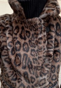 Manteau en fausse fourrure imprimé léopard avec un col haut en maille noire côtelée, doté de boutons noirs et d'une ceinture à la taille. Texture douce et tons bruns riches.