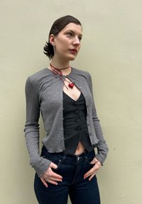 Cardigan cinza com textura canelada, de corte cropped, usado sobre uma blusa preta ajustada com botões. Completo com calças de ganga escuras e um colar em forma de coração vermelho.