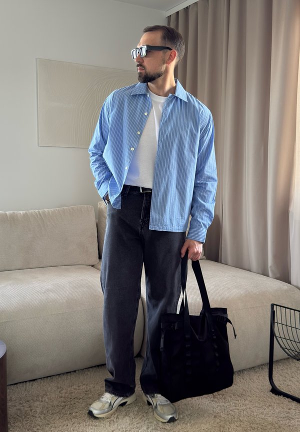 Homme portant des lunettes de soleil et une chemise rayée bleue tient un sac noir debout dans un salon avec un canapé beige et des rideaux.
