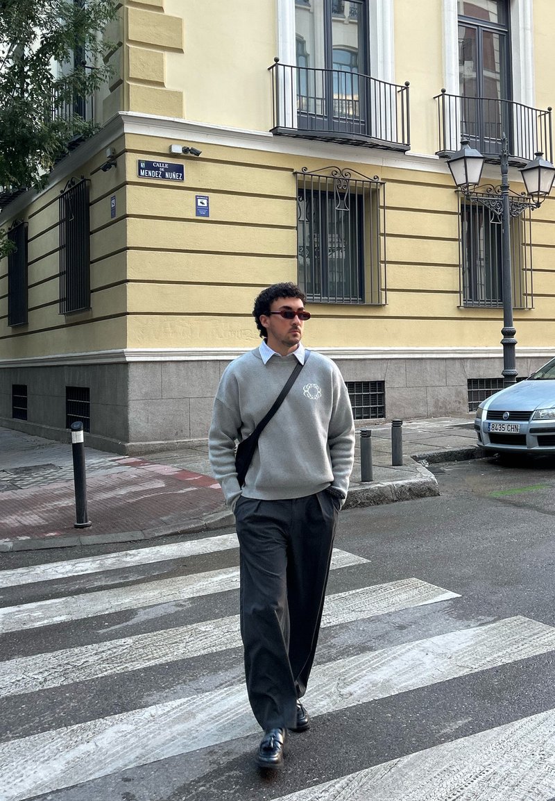 Pull gris avec logo, chemise à col blanc, pantalon sombre et chaussures noires. Marchant sur un passage pour piétons à côté d'un bâtiment jaune et de panneaux de signalisation.