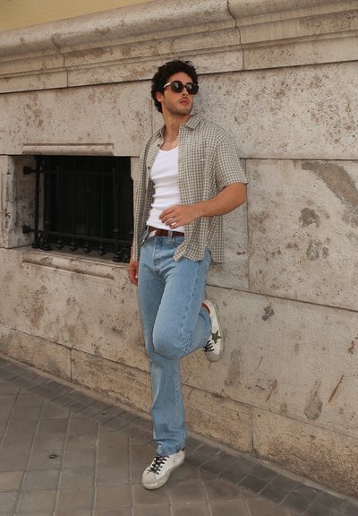 Camisa de cuadros de manga corta sobre una camiseta sin mangas blanca, jeans de color azul claro y zapatillas blancas con acentos verdes, apoyado contra una pared texturizada.