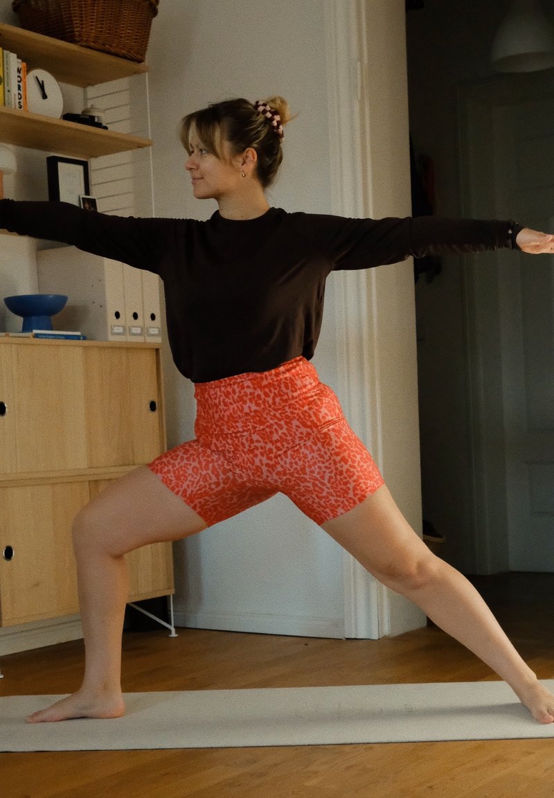 Femme pratiquant le yoga en posture du guerrier sur un tapis à l'intérieur, portant un haut noir à manches longues et un short orange à motifs, regardant vers la gauche.