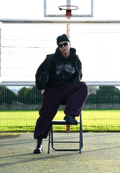 Sudadera negra con estampado gráfico, superpuesta a una chaqueta oscura, pantalones cargo morados holgados y botas negras. Sentado en una silla de metal, con un aro de baloncesto de fondo.