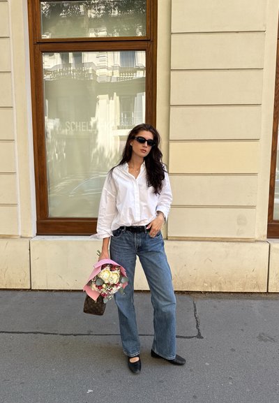 Camisa blanca de botones, jeans de pierna ancha de color azul claro, manoletinas negras, sosteniendo un ramo envuelto en papel rosa, con un bolso marrón texturizado.