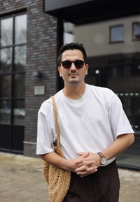 Vit löst sittande T-shirt, bruna byxor med dragsko, flätad halmning, runda solglasögon och en klocka med brunt läderarmband. Tegelvägg i bakgrunden.