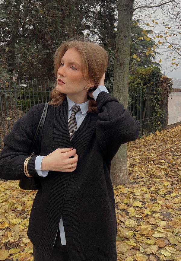 Zwarte gebreide cardigan met een sjaalkraag over een lichtblauw hemd en gestreepte das. Het model houdt een kleine zwarte handtas vast. Herfstbladeren verspreid over de grond.