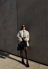 Grijze oversized blazer, wit shirt, zwarte stropdas, zwarte minirok, doorschijnende zwarte panty's, zwarte hoge hakken, koffiekopje en handtas vasthoudend.