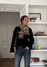 Femme portant un pull noir, une écharpe à carreaux, un jean et un sac noir, debout à l'intérieur près d'étagères blanches avec des livres, de la décoration et des flacons de parfum.