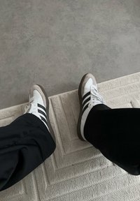 Pieds portant des baskets blanches à rayures noires, debout sur un tapis beige texturé à côté d'un sol gris lisse.