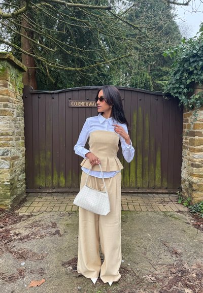Mujer con pantalones anchos beige y top estilo corsé sobre una camisa blanca, sosteniendo un bolso blanco, parada frente a una puerta marrón con un cartel que dice "CORNERWAY".