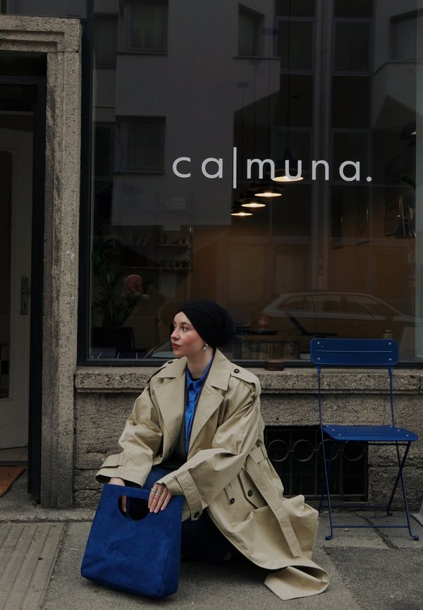 Mujer con gabardina beige y pañuelo negro en la cabeza se agacha fuera de una cafetería con una silla azul, sosteniendo un bolso azul a juego en la acera.