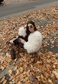 Pihe-puha fehér pulóver, sötét nadrág és napszemüveg. Barna és narancssárga őszi levelekkel borított talajon ül, barna táskát tartva.