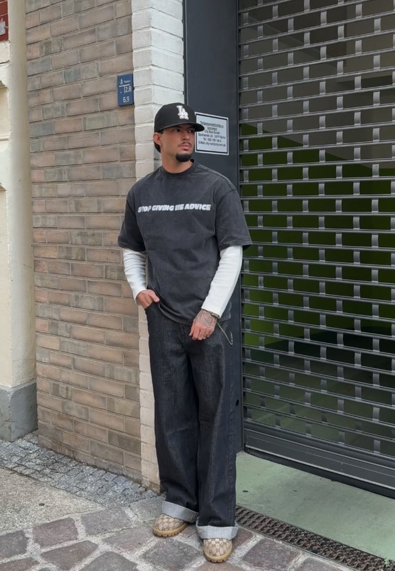 Μαύρο μπλουζάκι με λευκό κείμενο, επικαλυμμένο πάνω από ένα λευκό μακρυμάνικο πουκάμισο. Σκούρα παντελόνια wide-leg, παπούτσια σε ταμπά με σχέδιο, στέκεται μπροστά από φόντο με υφή.