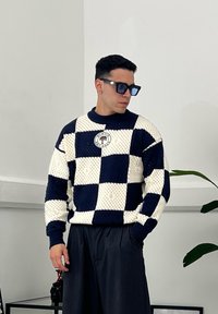 Gebreide trui met een marineblauw en crèmekleurig dambordpatroon en een ronde logo patch. Gekleed met zwarte wijde broek.