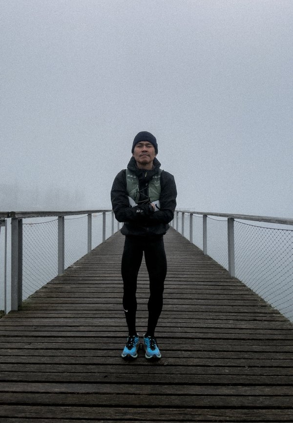 Hombre vestido con ropa deportiva negra y zapatillas de correr azules, de pie con los brazos cruzados en un muelle de madera con niebla y barandillas de metal.
