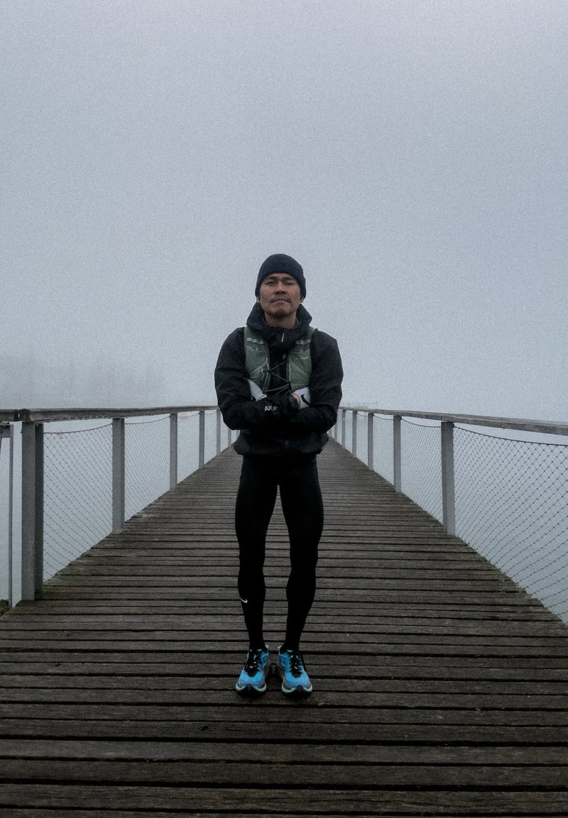 Άνδρας με μαύρα αθλητικά ρούχα και μπλε παπούτσια τρεξίματος στέκεται με τα χέρια σταυρωμένα σε ομιχλώδη ξύλινη προβλήτα με μεταλλικές κουπαστές.