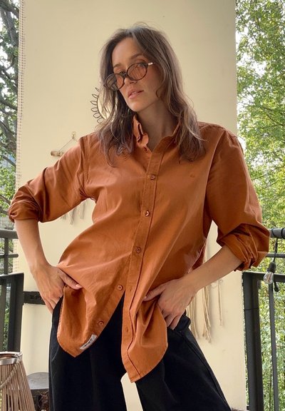 Camisa marrón de botones con mangas enrolladas, hecha de una tela suave. Presenta un logotipo sutil y contrasta con pantalones negros holgados.