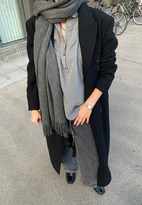 Juodas vilnonis paltas ant pilkai dryžuoto marškinėlio, pilkas šalikėlis su kutais, tamsūs džinsai ir juodi blizgantys batai. Matomi sidabrinis laikrodis ir žiedai.