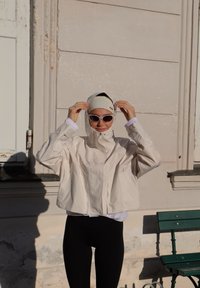 Veste beige courte avec une capuche, texture lisse, fermeture éclair à l'avant et deux poches latérales ; portée avec des leggings noirs et des lunettes de soleil.