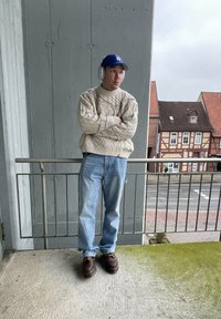 Pull beige en tricot, jean bleu clair, chaussures marron foncé, écouteurs blancs, casquette bleue, debout sur un balcon avec des bâtiments en arrière-plan.