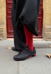 Svarte Nike-sneakers med teksturert overdel, røde aksenter og synlig luftpolstring. Kombinert med ribbestrikkede røde sokker og løse svarte bukser.