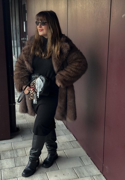 Abrigo de piel sintética marrón, suéter de cuello alto negro, falda midi negra, botas altas negras; bolso con un charm de animal, gafas de sol redondas.